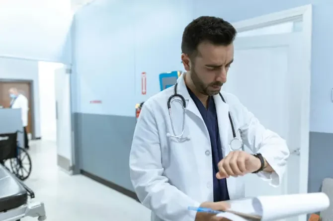 A doctor in a white coat with a stethoscope around their neck enters a modern hospital while checking their wristwatch, illustrating time management in healthcare.